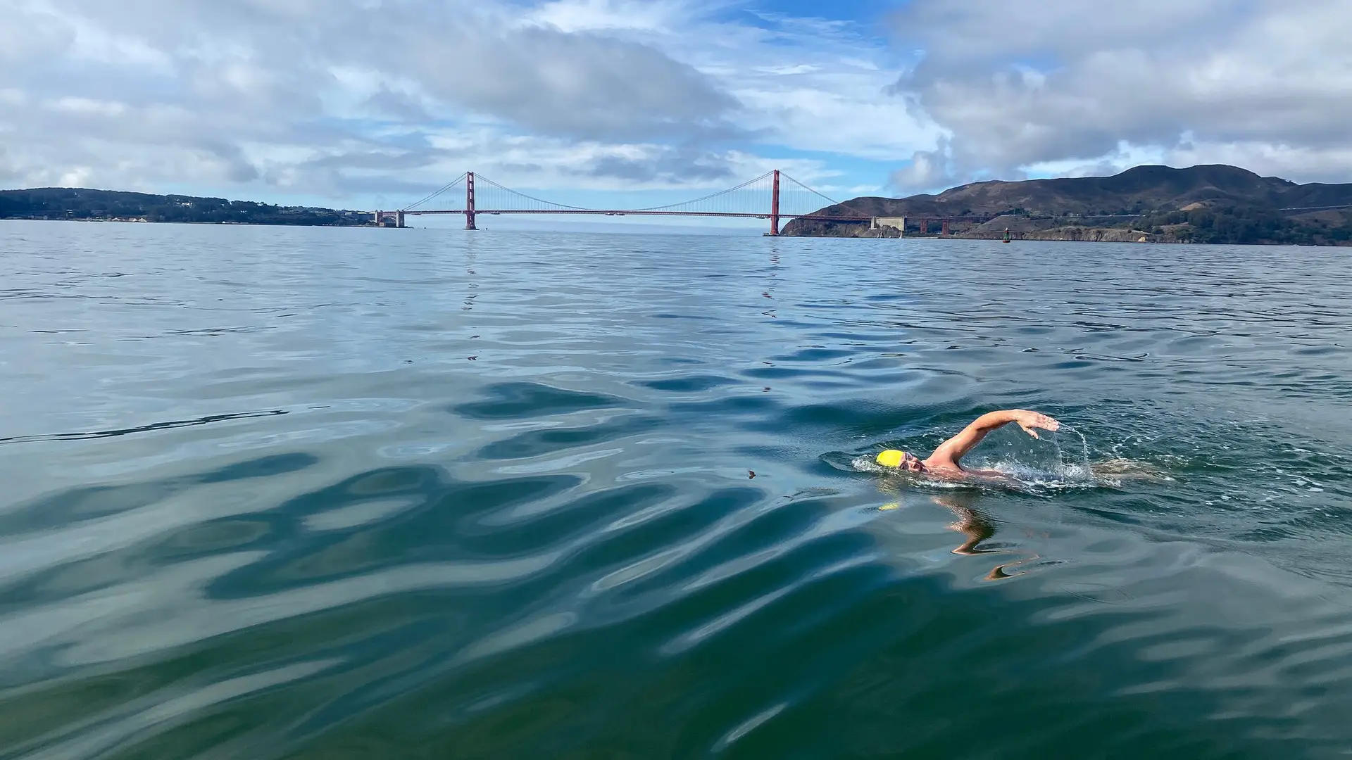 A solo open water swimmer wearing a yellow swim cap swims freestyle across San Francisco Bay with the Golden Gate Bridge visible in the background under a partly cloudy sky.
